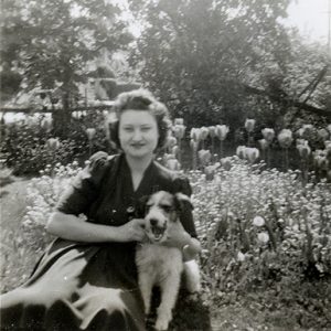 Black and white image of a woman in a dress seated in a yard with flowers, holding a dog.