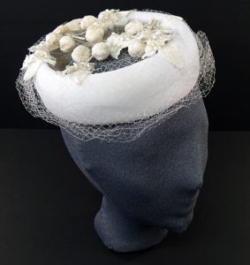 Head of a mannequin is seen sporting a white pillbox hat with white netting and a floral motif. Head is tilted at a 3/4 angle.