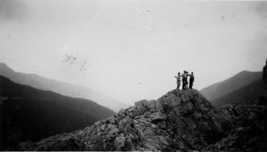 Black and white image of 4 figures standing atop a rocky mountain ride, the background shows the silhouette of mountain peaks in the valley.