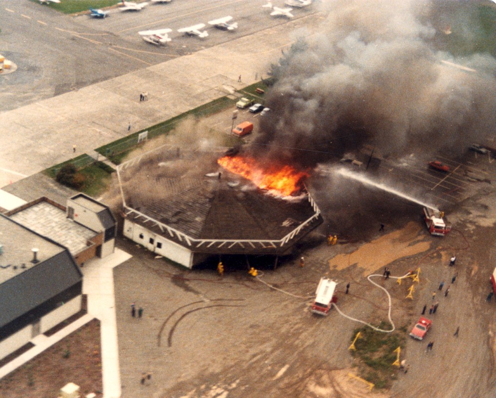 Photograph consists of a low elevation aerial view of the airplane hangar on fire at the Chilliwack Airport.  Image shows two firetrucks and numerous firefighters battling the blaze, 29 October 1986. [P5644] 