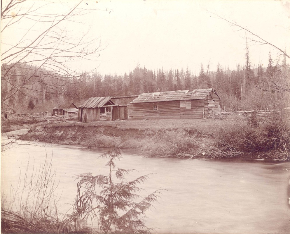 Photograph consists of a view of the longhouse at Skowkale First Nation with the Ts'elxwéyewq River (Chilliwack) in the foreground, ca. 1890s.  [P5562]